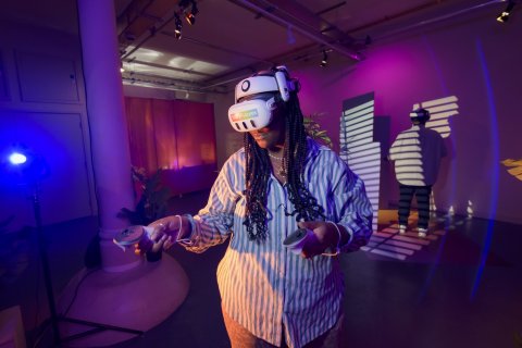 A young Black woman with long braids stands in a headset engaging in a mixed reality experience. 