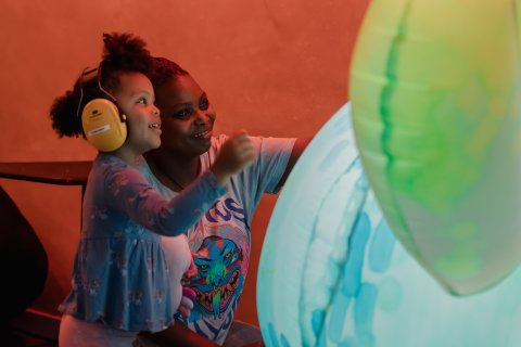 A young brown skinned child is help by her smiling mother and they are both touching a giant inflatable robotic plant.