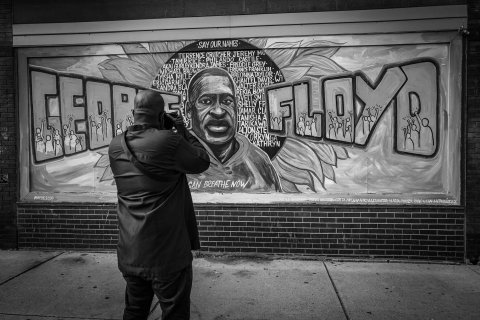 A person stands photographing a colorful mural honoring George Floyd, surrounded by names of other victims of racial violence.
