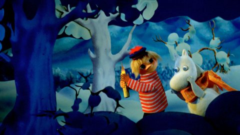 A boy in a striped sweater holding a candle, stands beside a startled Moomin, amid a snowy landscape with dark trees.