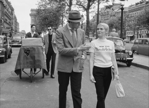 A man in a suit gestures while walking alongside a woman in a "New York Herald Tribune" t-shirt on a busy street with vintage cars.