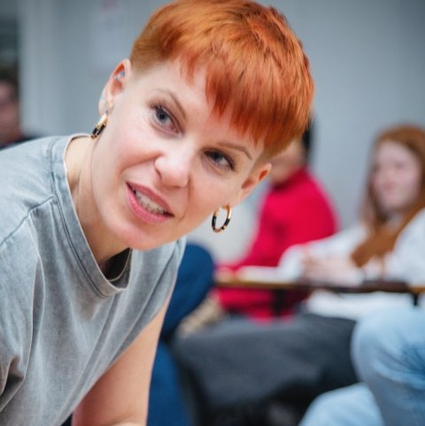 Gemma is a young white woman with red hair and hoopearrings and is peering off to the left.