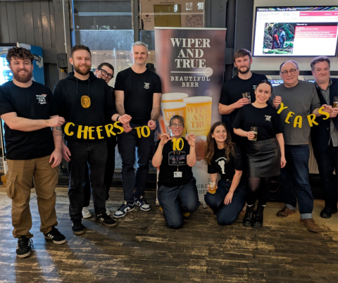 A picture of people standing in front of a Wiper and True banner holding a sign saying 'Cheers to 10 years'