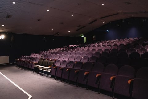 Empty dimly lit cinema auditorium with rows of purple cushioned seats and a carpeted aisle on the left side.