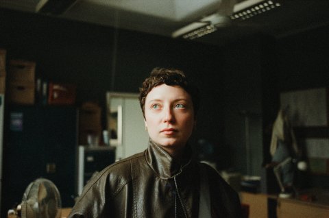 Thea Gajic headshot. A woman with short hair looks thoughtfully to the side in a dimly lit room. They're wearing a leather jacket, surrounded by shelves and boxes.