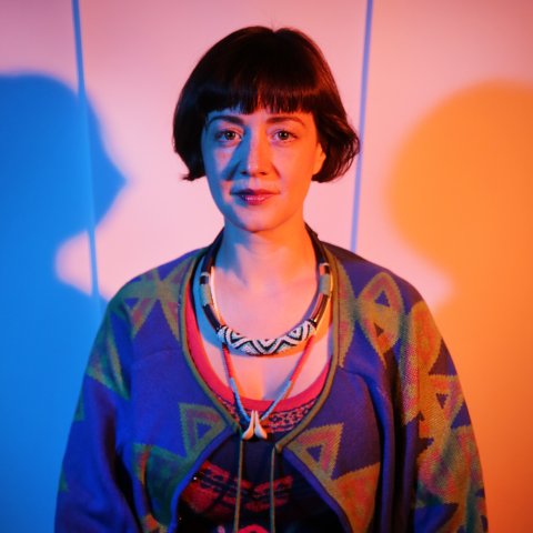 A woman with short hair stands against a wall lit with blue and orange hues. She wears a patterned sweater and a decorative necklace, exuding a calm and artistic vibe.