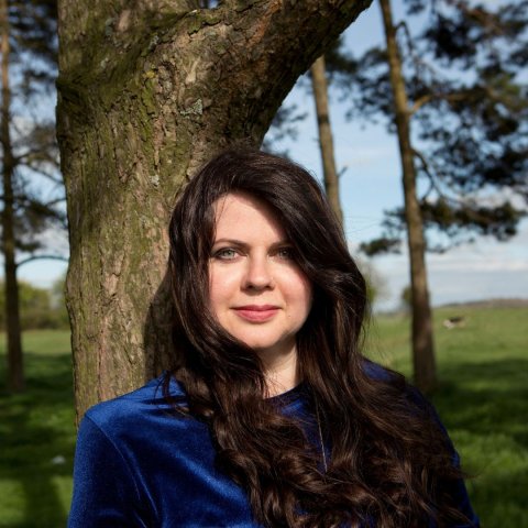 A woman with long dark hair in a blue top stands by a tree in a sunlit forest clearing. The scene feels serene and natural, with a clear blue sky.