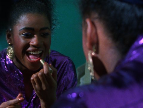 Person wearing a shiny purple outfit and gold earrings, looking into a mirror against a teal background.