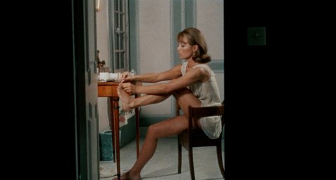 Woman in a white slip sitting on a chair at a vanity table, touching her toes in a softly lit room with blue-trimmed walls.