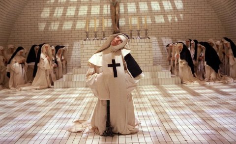 Nuns in white and black habits gathered in a tiled chapel, one nun in front with a large cross looks upward beneath a crucifix.