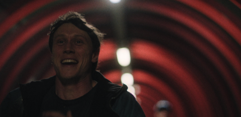 A man with a joyful expression runs through a red, tunnel-like structure illuminated by circular lights.