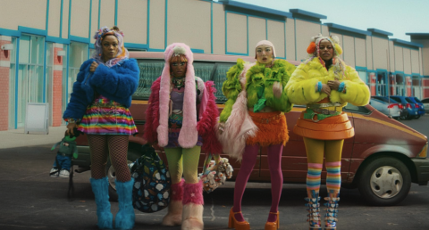 Four women in hyper-colourful outfit stand in front of a car in a car park.