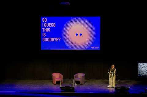 A speaker stands at a lectern on a dark theater stage beneath a blue screen reading “SO I GUESS THIS IS GOODBYE?”, with two chairs.