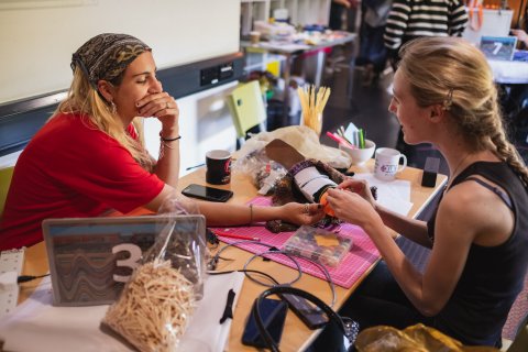 Two women collaborate at a crowded craft table, examining a small handmade item amid beads and tools; their focused, relaxed workshop scene feels creative and welcoming.