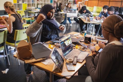 Students collaborate on laptops and electronics at crowded classroom tables, sharing ideas in a lively, focused workshop led by hands-on learning.