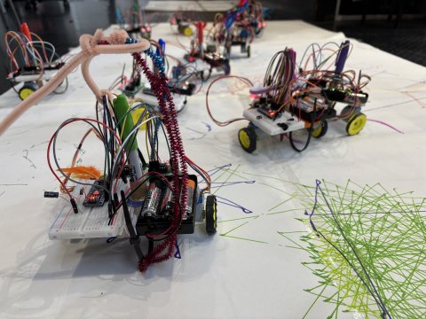 Small experimental robots with exposed wires and colorful sensors sit on a white floor, surrounded by tangled green and pink lines, creating a playful, inventive lab scene.