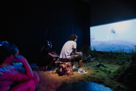 A small group sits in a dark, grass-covered gallery, watching a large screen show a misty blue landscape; the immersive installation feels quiet and contemplative.