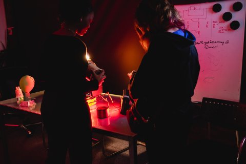 Two people in dark clothing study a glowing red-lit tabletop beside a whiteboard covered in sketches and notes, creating a moody, focused lab scene.
