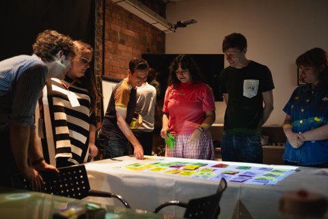Six young adults gather around a table to study colorful printed designs in a dim art studio, sharing a focused, collaborative moment.