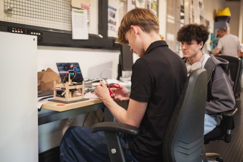Two students collaborate at an engineering workshop desk, assembling a small model with tools beside a laptop; the scene feels focused and hands-on.