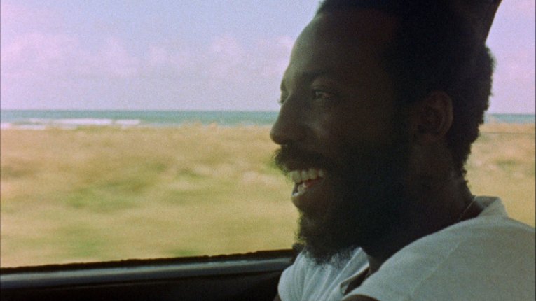 Photo of a man sitting by the  open window of a moving car, from the 70s in Jamaica