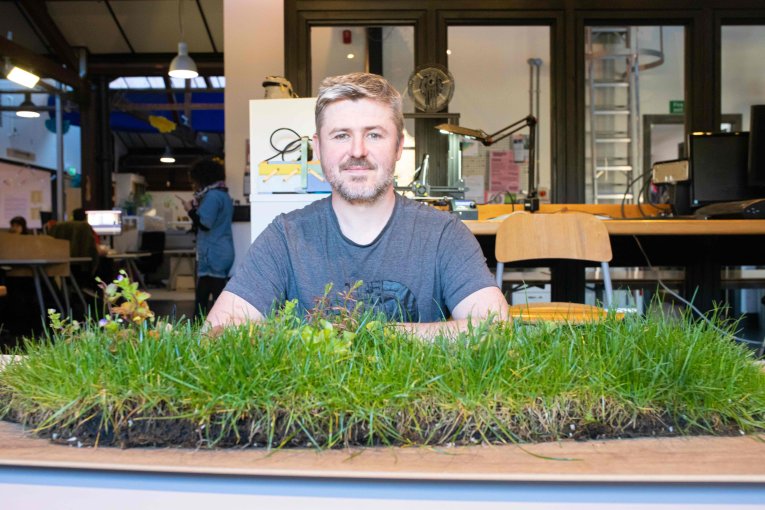 A man in a blue tee smiles directly at the camera. He sits behind a table, on it - a lush layer of turf.