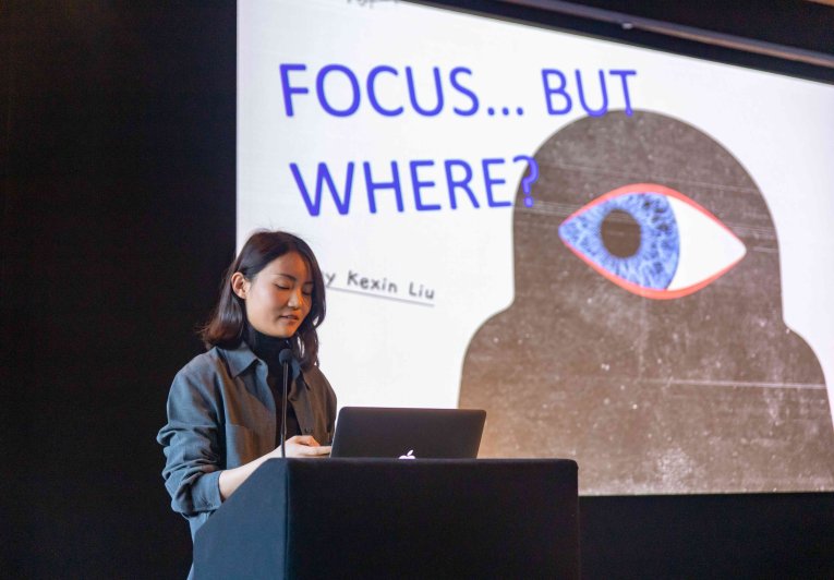 A woman stands before a slideshow, projected onto the wall behind her. The slide reads, 'Focus...but where?'