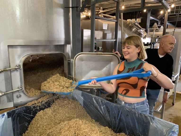 Someone shovels brown hops into a silver machine, a smile on their face.