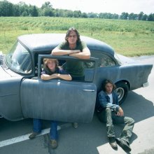 Film still of three people with a parked car, one standing, one in the fron seat and one lying on the road leaning against the car