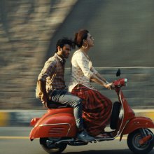 Two people ride on a red scooter, the background a blur.
