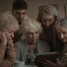 A group of four older women and 30-something man crowd around a tablet