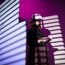 A woman stands with a VR headset on and controllers in here hand, the background is purple and pink with various shadows.