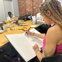 A person with braided hair sketches in a large notebook during a meeting at a wooden table with food and drinks present.