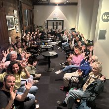 hoto of Deaf Conversations About Cinema discussion group at Watershed. A large group of people sitting in a long corridor together - waving their hands in the air after the discussion.