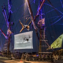 Celluloid Sail - tall ship docked to harbourside with projections on sails and acrobat hanging from rope.