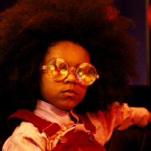 A child wears large, round mirrored sunglasses and dungarees and looks towards the camera. 