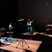 A group of musicians in a dark studio. One plays guitar, one drums and a piano in the background.