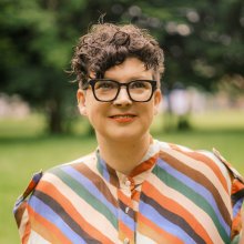 Head and shoulders of a woman with short hair and glasses wearing a multi coloured, stripey top with trees in background.