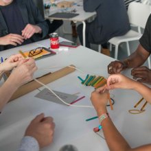 A diverse group if people are doing an activity together on a white tables with string and colourful lollipop sticks.