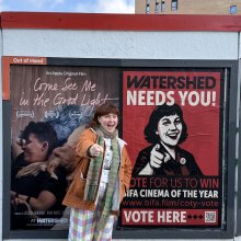 An outdoor poster site - a person stands to the side smiling and pointing in the same pose as the person in the poster which also has the words' Watershed Nees