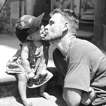 A black and white photo of the artist Andrew Kötting kissing his daughter Eden on the cheek.