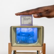 A hand hovers over a small screen atop a retro TV showing a blue image. The screen displays text with words about a "palace" and a "lake." The vibe is artistic and thought-provoking.