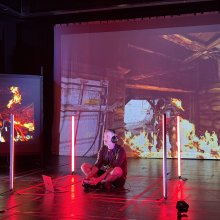 A person sits cross-legged on the floor, wearing headphones, surrounded by vertical red lights. Intense video game scenes with fire project in the background.