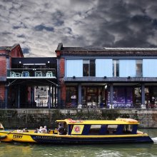 exterior shot of the Watershed building, including the balcony, there is a yellow ferry on the water passing the building and the sky is dark and stormy.