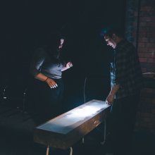 Two people stand in a dimly lit room, focused on a glowing rectangular table. The scene conveys curiosity and concentration against a brick wall backdrop.