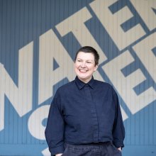 A smiling woman stands outside the Watershed blue panel - she has short cropped hair, and is look away from the camera.