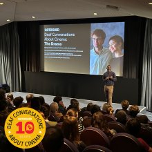 Inside a full cinema, man at the front of the audience introduces a Descriptive Subtitled screening of The Drama
