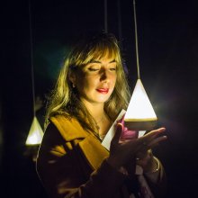 A woman holds her hand up to a glowing triangular light. It illuminates her face in a dark room.