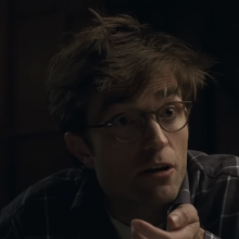 A person gestures while sitting at a table in a dimly lit room, with a warm light source in the background.