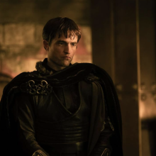 A person dressed in dark armour and cloak stands in a dimly lit room with stone walls and warm lighting.